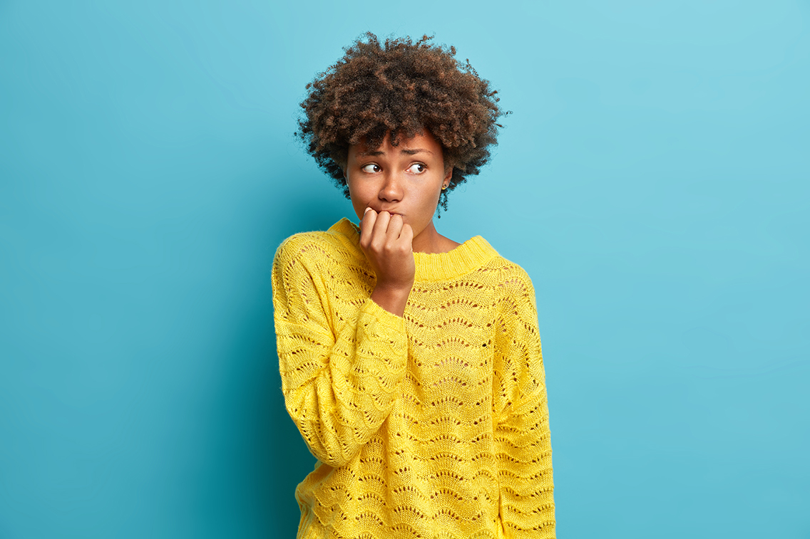 Portrait of nervous Afro American woman keeps hands near mouth feels concerned before important interview hesitates about something dressed in knitted yellow jumper poses against blue studio wall